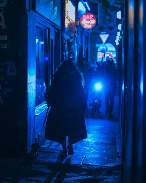 Person walking a dog through a narrow alley at night, illuminated by blue neon lights.