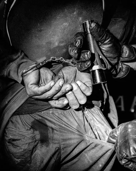 Close-up of a handcuffed person and another person searching his pants pocket with a flashlight.
