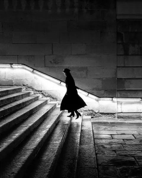 A woman in a long coat is walking up a staircase at night. The banister is illuminated.