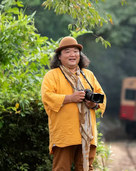 Man with camera stands beside railroad tracks and a train in background.