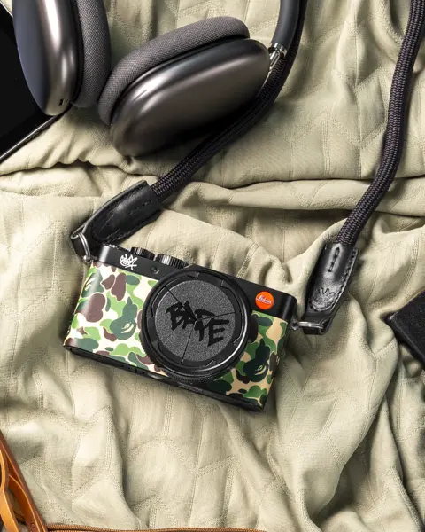 A camera, tablet, headphones, sunglasses, perfume and wallet laying on a bedcover.