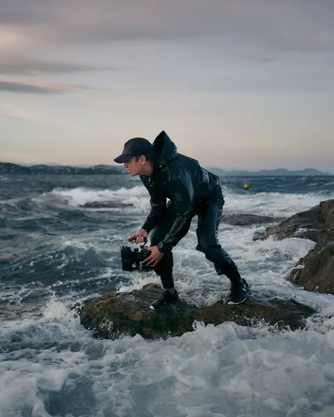 A person is standing on a rock in a stormy sea, holding a pair of binoculars.