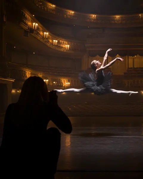 A person in the shade takes a picture of a ballerina jumping in an illuminated theatre.