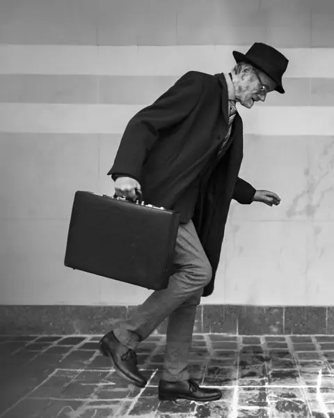 Elderly man in suit with hat and briefcase walking on icy cobblestones.