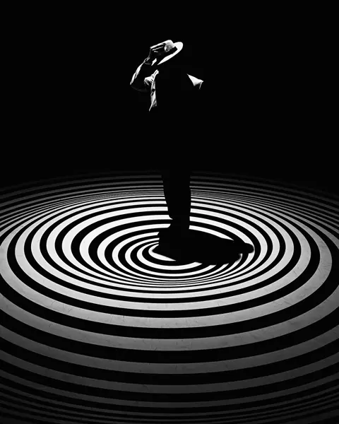 A single person with a hat stands in the centre of concentric circles on a dark background