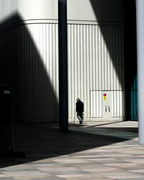 An old man walks in front of a building with shadows beside him.