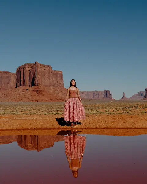 Woman standing in a desert landscape with red rock formations in the background, the image is reflected in the water in front of her