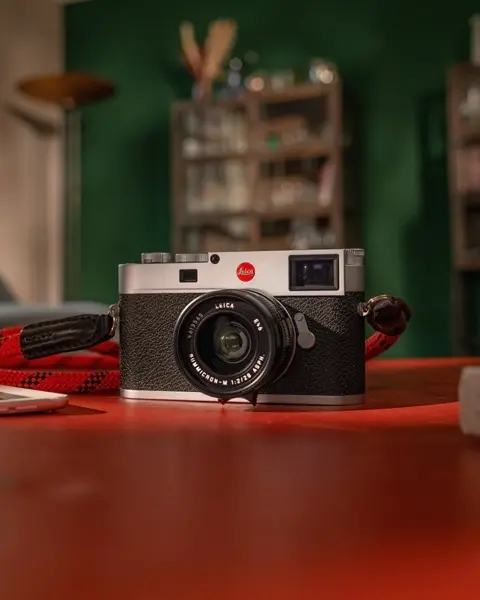 Leica camera with red strap on a table, blurred background with sofa and bookshelves.