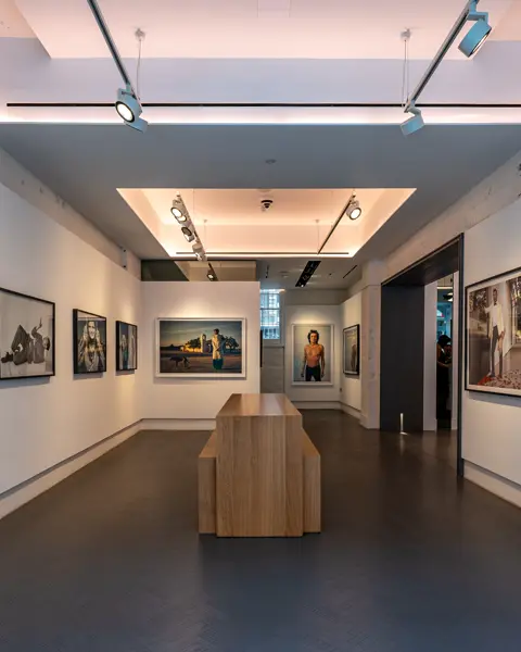 Exhibition room with photographs of various people and a wooden counter and benches in the centre of the room.