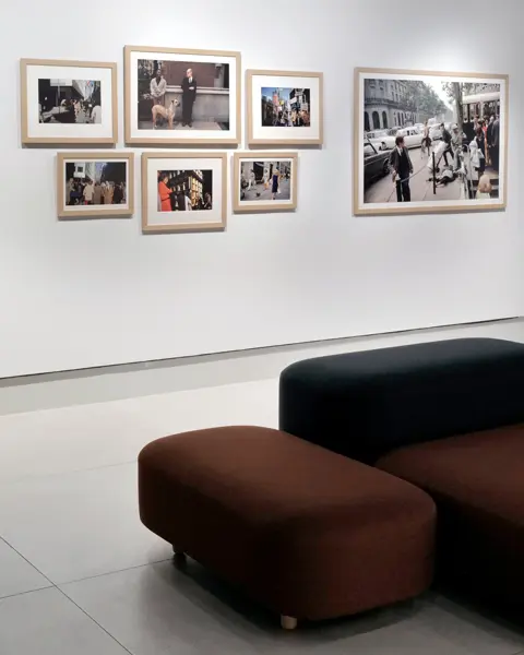 Framed pictures of people in the city hang on a white wall, with black and red upholstered stools in front of them.