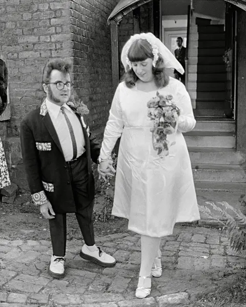 A wedding couple and a woman in a dress stand in front of a building.