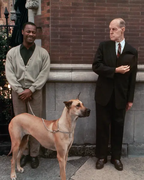 Two men, one with a dog on a lead, stand in front of a brick wall.