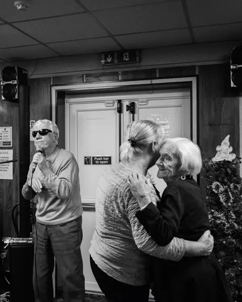 A man sings into a microphone while two older women dance and a man plays the keyboard in the background.