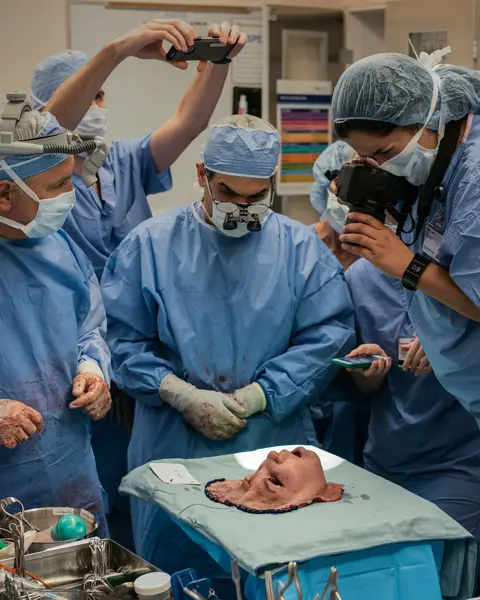 A group of people in surgical gowns look at a mask representing a face, and some take pictures of it.