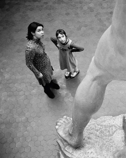 Two people stand in front of a large statue and look upwards.