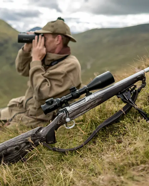 A man sitting on a hill with binoculars. In front of him is the Leica Magnus riflescope mounted on a weapon