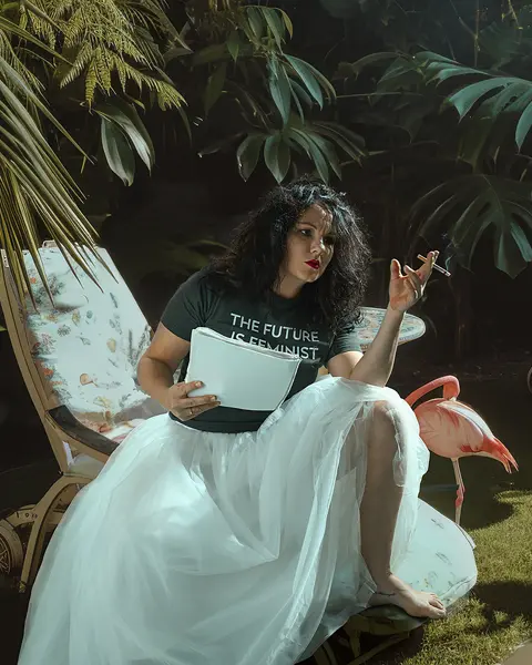 Woman with white tulle skirt and cigarette in her hand on a lounger with flamingos standing around her