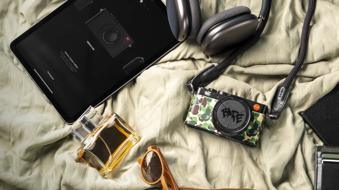 A camera, tablet, headphones, sunglasses, perfume and wallet laying on a bedcover.