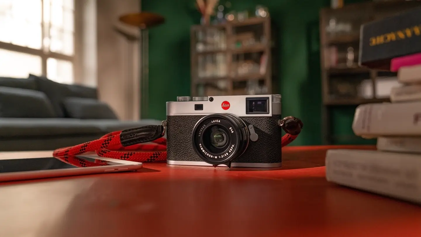 Leica camera with red strap on a table, blurred background with sofa and bookshelves.