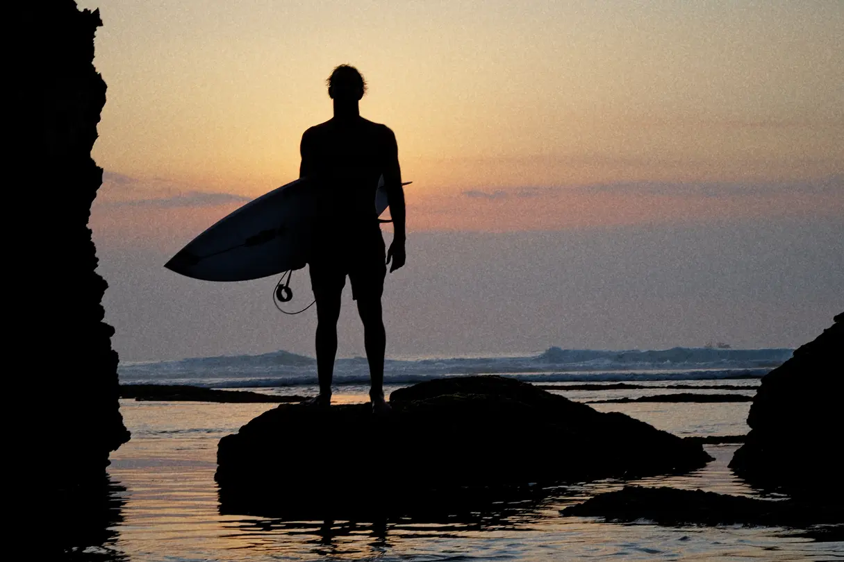 Surfer standing on a rock at sunset with his board under his arm