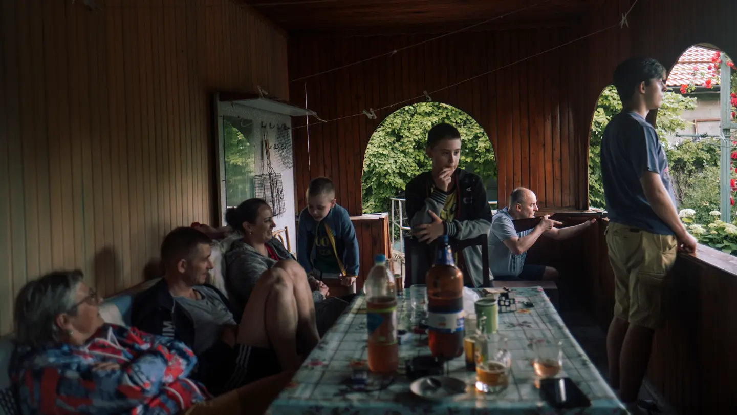 A big family is sitting at a table on a covered terrace and a man is holding a rifle aiming outside.