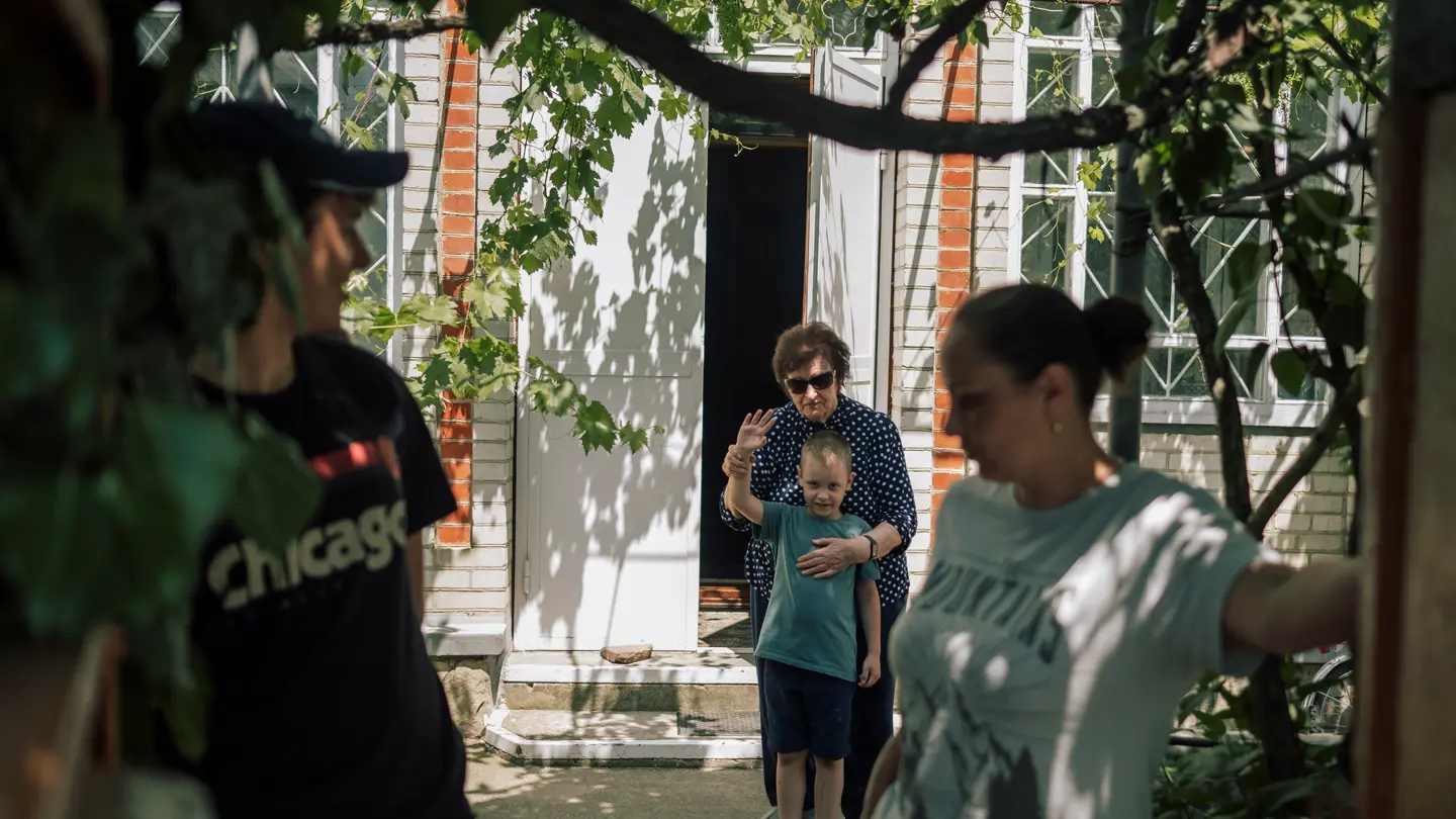 A grandmother stands behind her grandchild and waves to his parents with him.