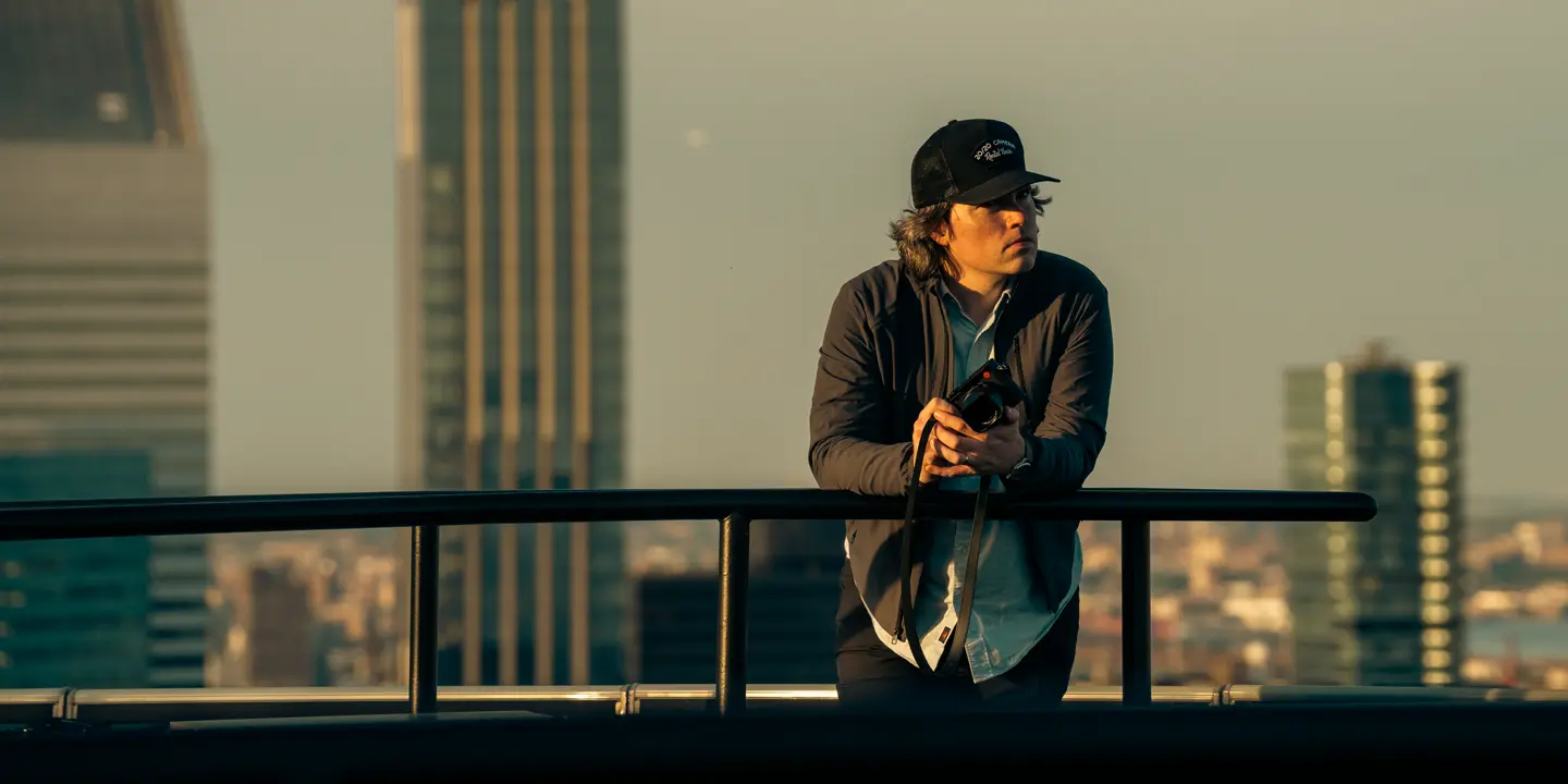 A man holding a camera leans against a railing with skyscrapers in the background.