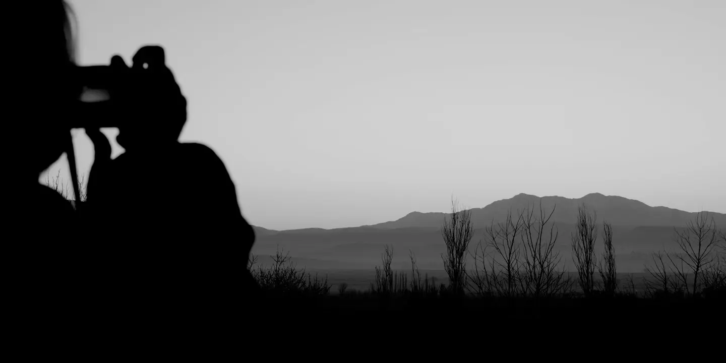 Black-and-white photograph of a person taking a picture of a lake and mountains in the background.