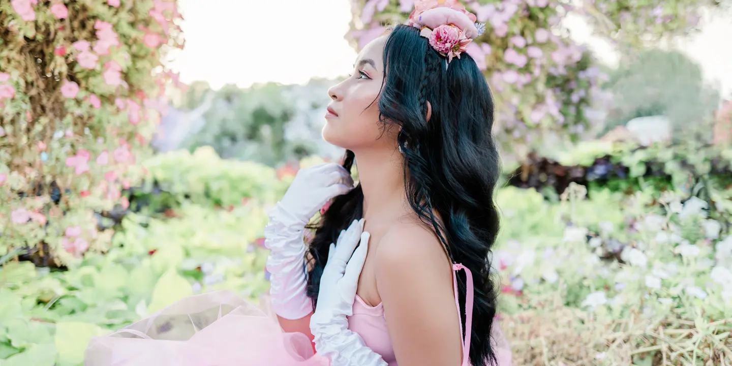Woman in a pink dress and white gloves is sitting in a rose garden looking to the side.
