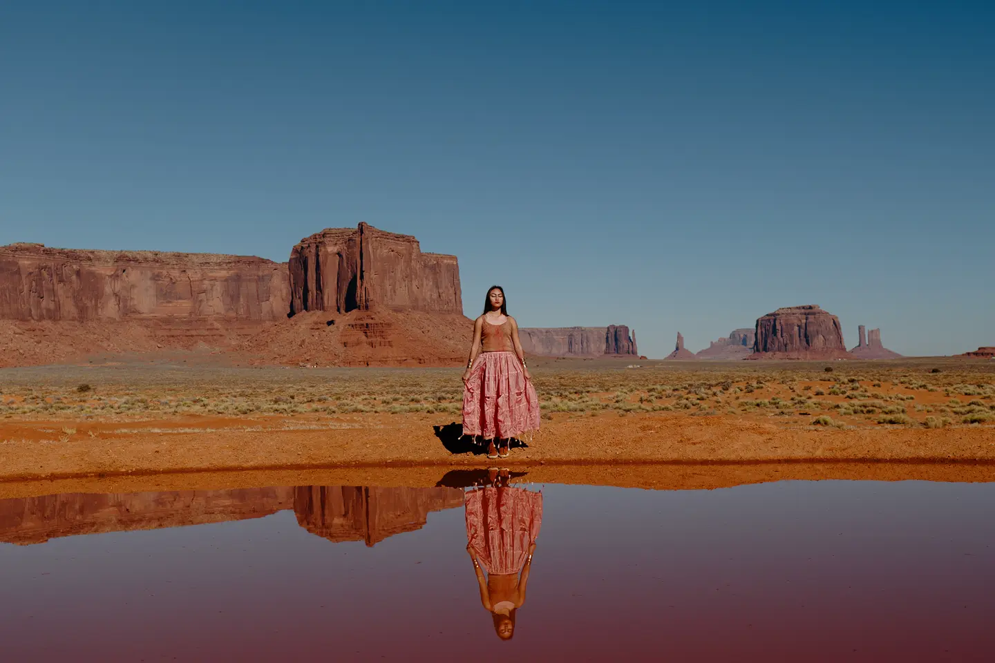 Woman standing in a desert landscape with red rock formations in the background, the image is reflected in the water in front of her
